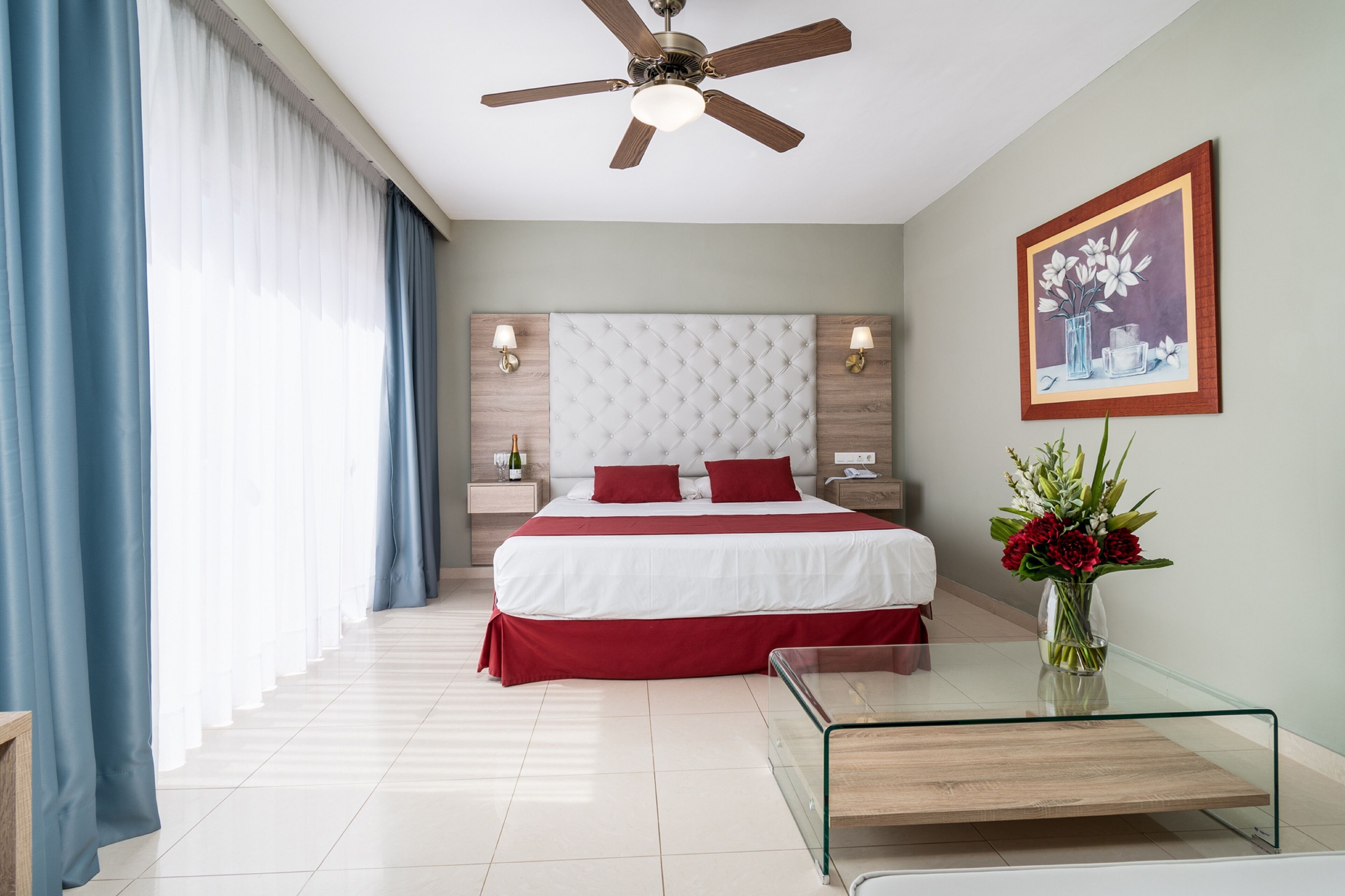 Una habitación de hotel moderna con una cama grande, una mesa de centro de vidrio, un ventilador de techo y un cuadro en la pared.