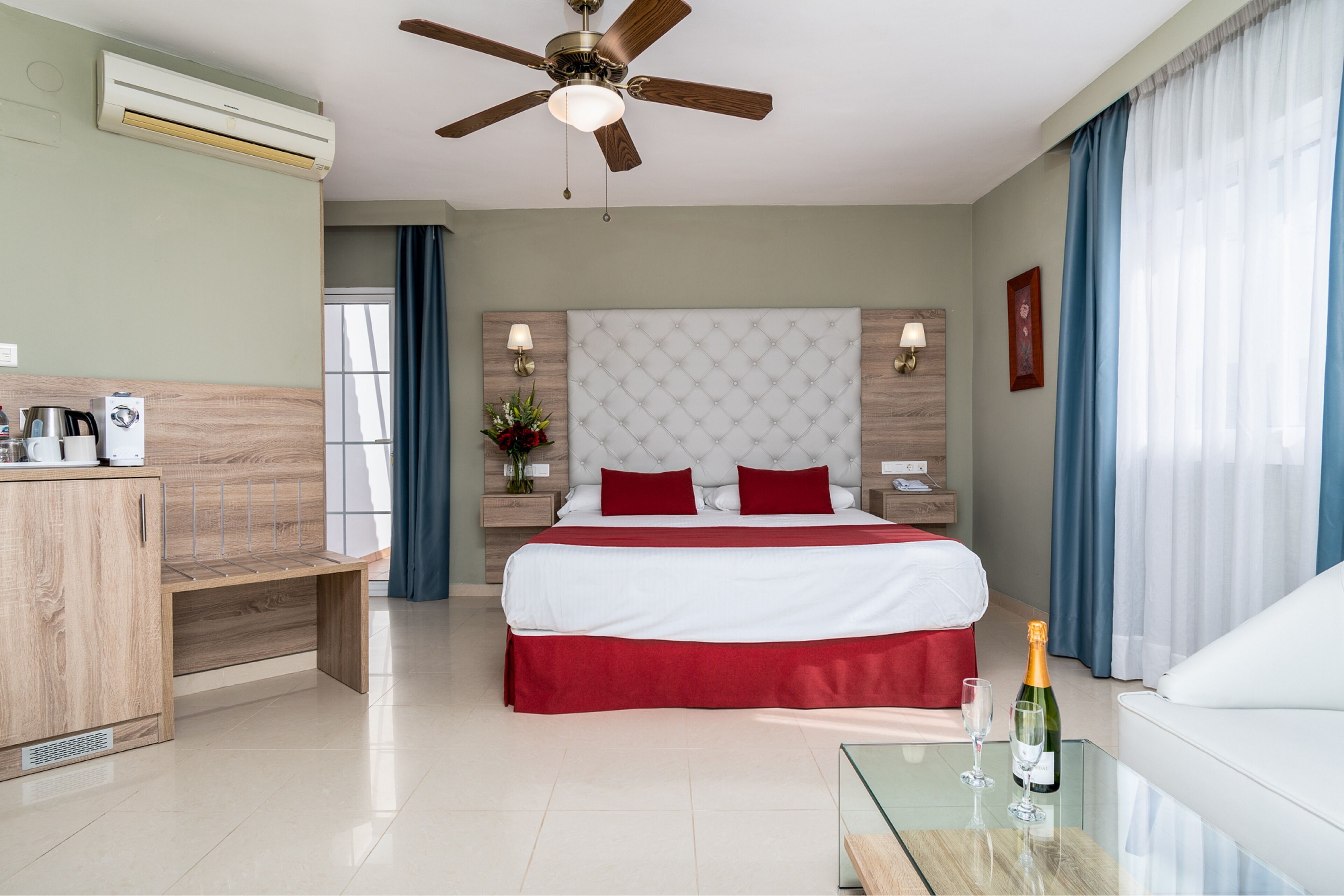 A hotel room with a bed, a glass table with a bottle and glasses, a wooden bench, a ceiling fan, and an air conditioning unit.