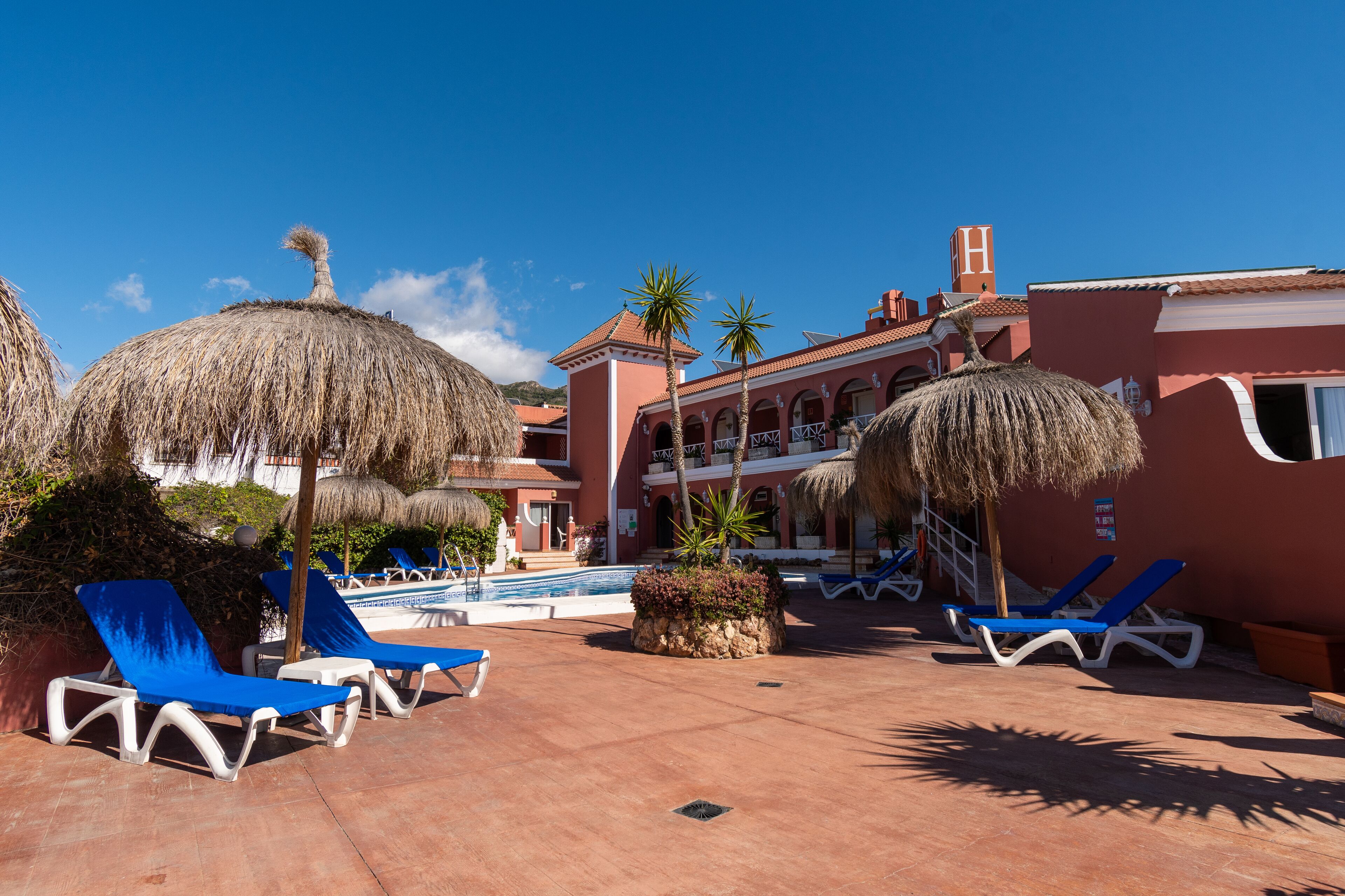 A resort pool area with lounge chairs and thatched umbrellas, a pool, and a building with a red facade and balconies.