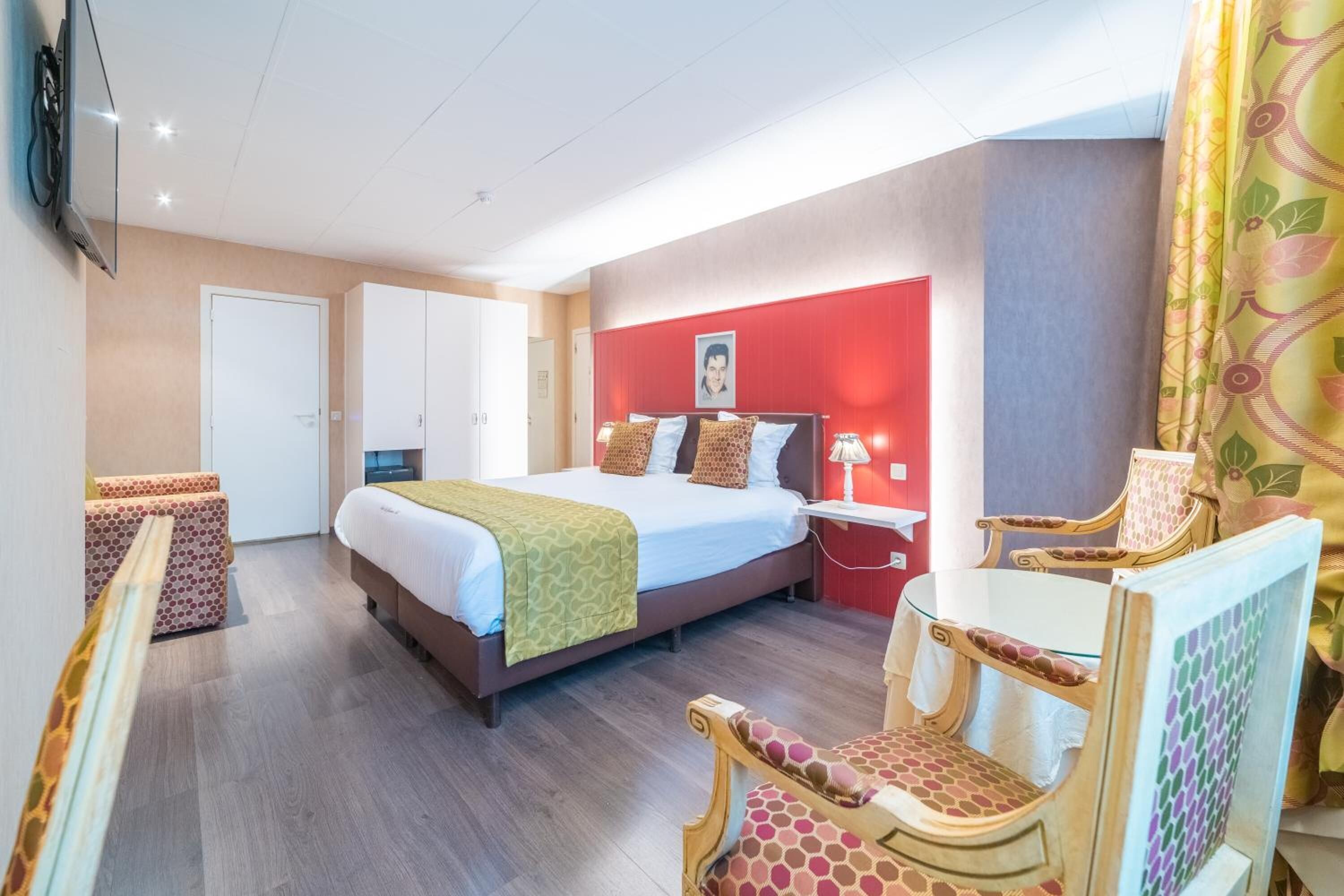 Habitación de hotel con cama, mesitas de noche, televisión, armario y zona de estar con sillones estampados.