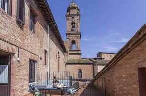 A courtyard with a brick building, a table with chairs, and a bell tower in the background. - Il Chiostro del Carmine (Siena)
