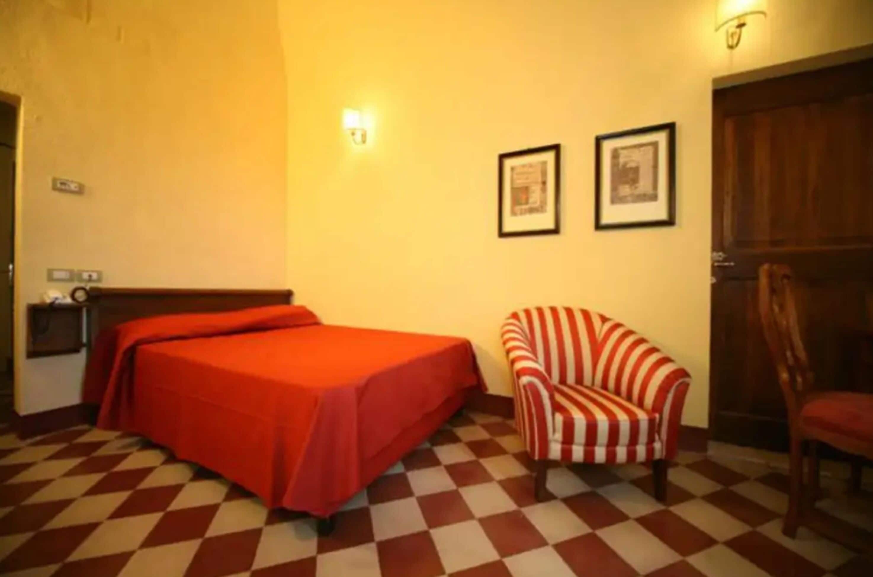 A room with a checkered floor, a bed with a red cover, a striped armchair, and two framed pictures on the wall.