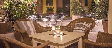 An outdoor seating area with wicker furniture and a table set for two, surrounded by potted plants and a brick building in the background.