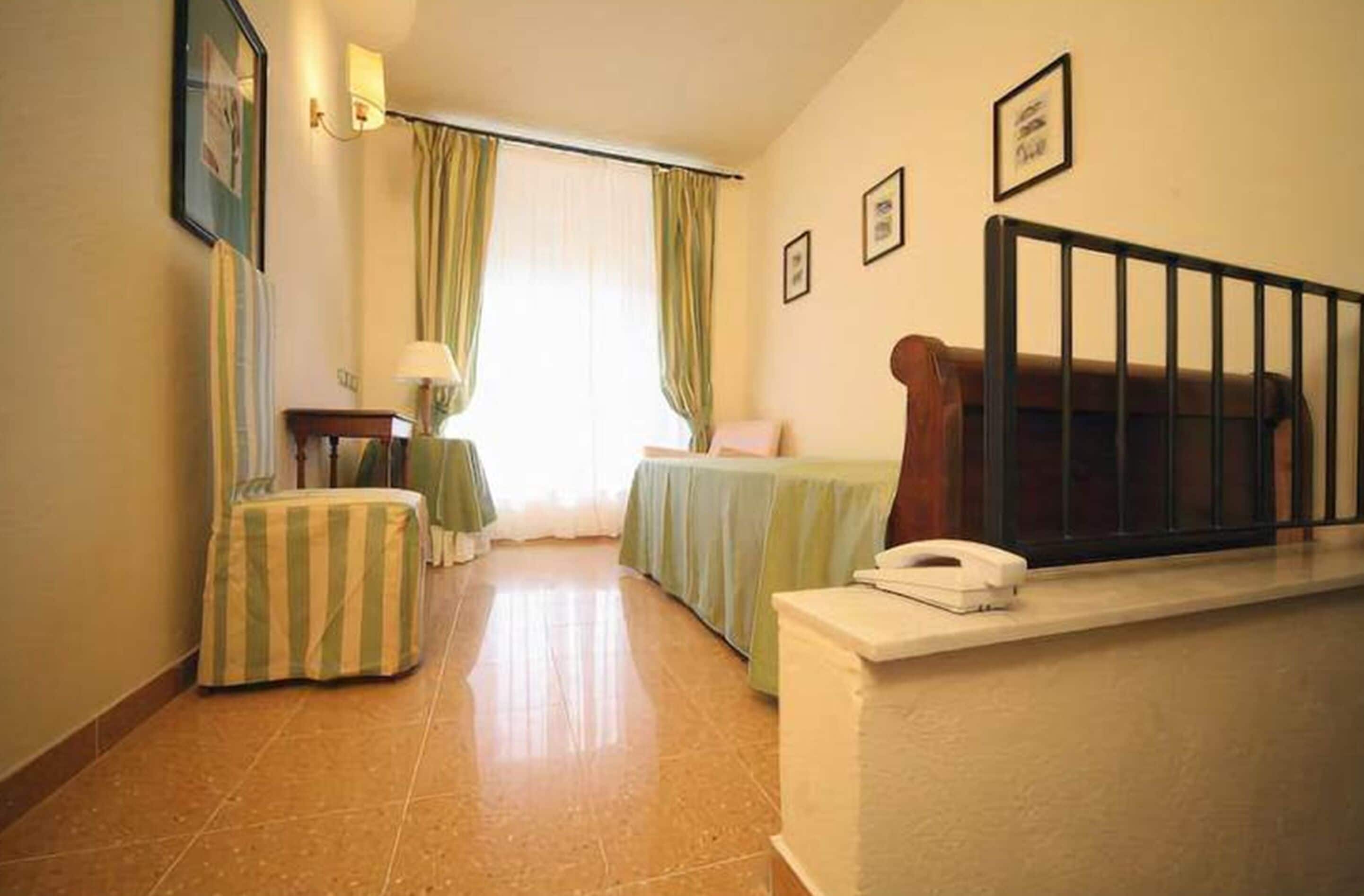A hotel room with a bed, a desk with a lamp, a telephone, a window with curtains, and a staircase railing.