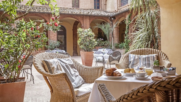 Un área de comedor al aire libre con mobiliario de mimbre, una mesa dispuesta con croissants, un tazón de fruta y un vaso de jugo de naranja.