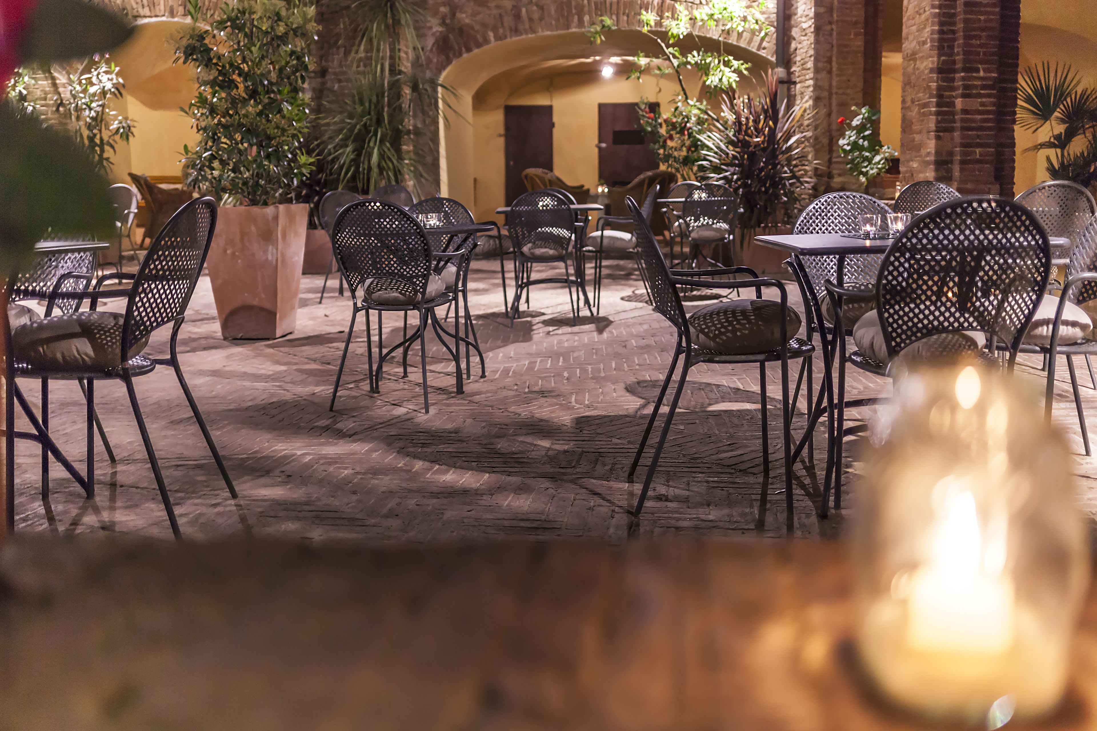 Un caffè all'aperto con sedie e tavoli in metallo, circondato da piante in vaso e un muro di mattoni.