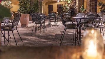 An outdoor café with metal chairs and tables, surrounded by potted plants and a brick wall.