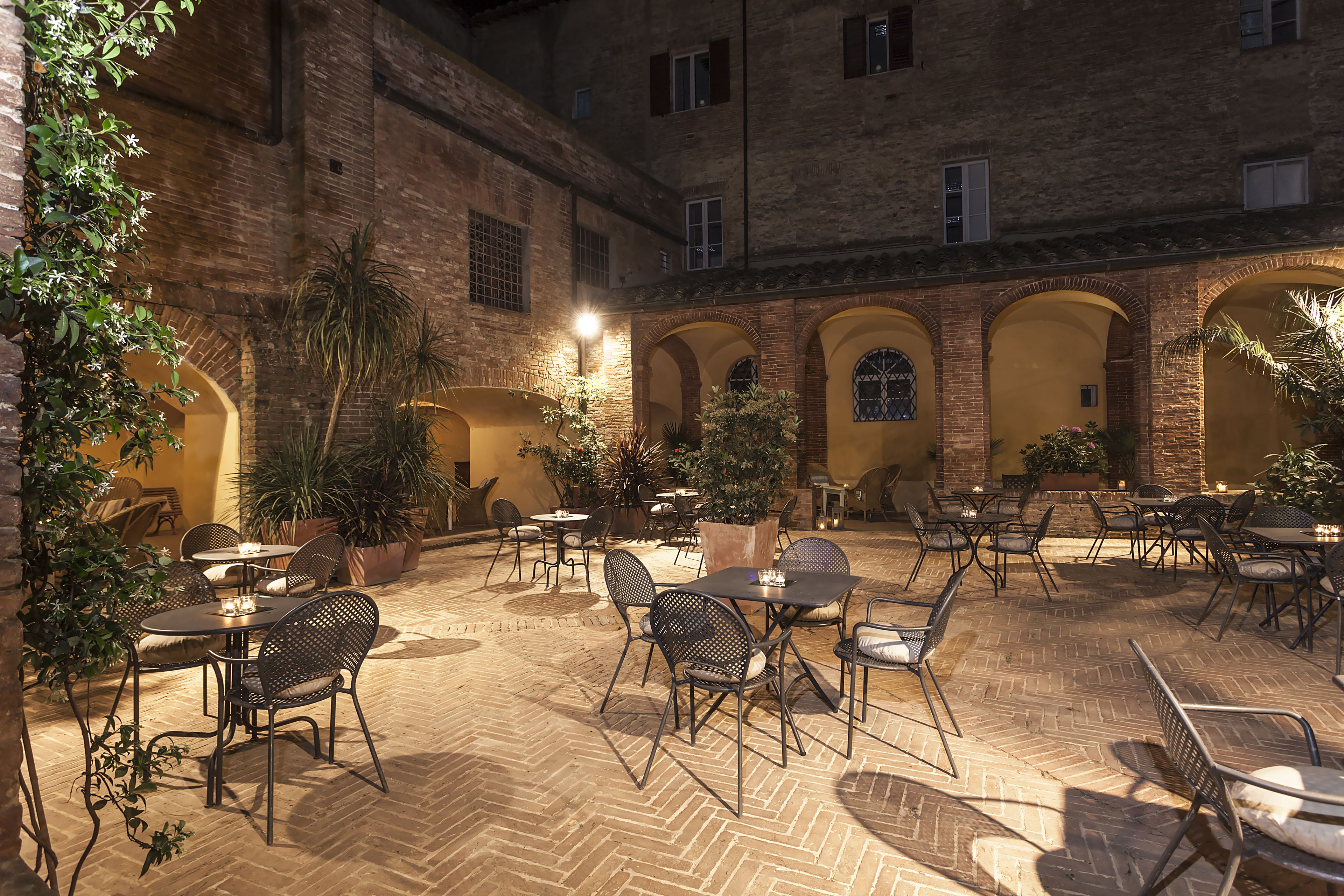 An outdoor patio area with brick flooring, multiple tables and chairs, potted plants, and a brick building in the background.
