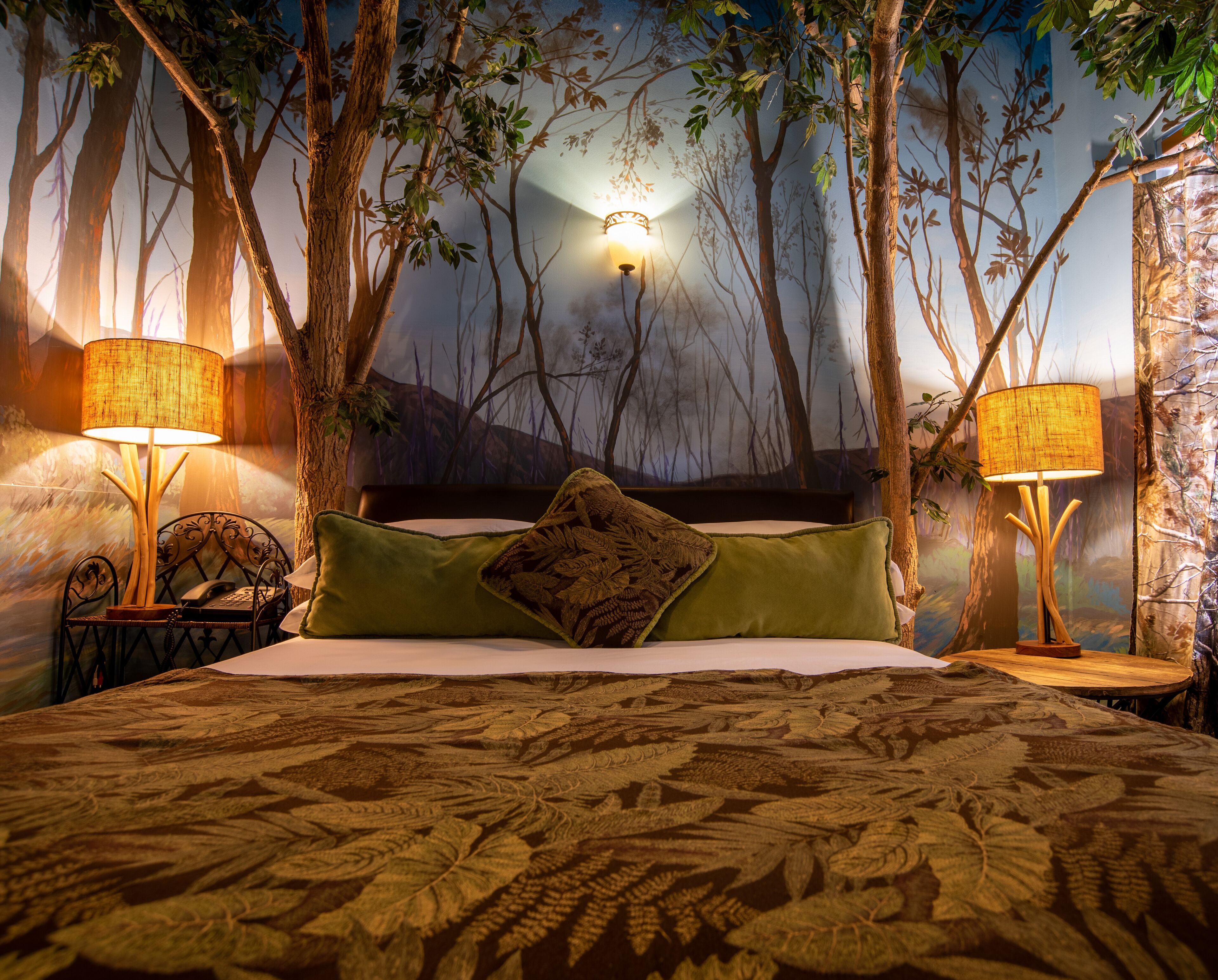 A bedroom with a forest-themed mural, a bed with a patterned bedspread, two bedside lamps, and a nightstand with a telephone.