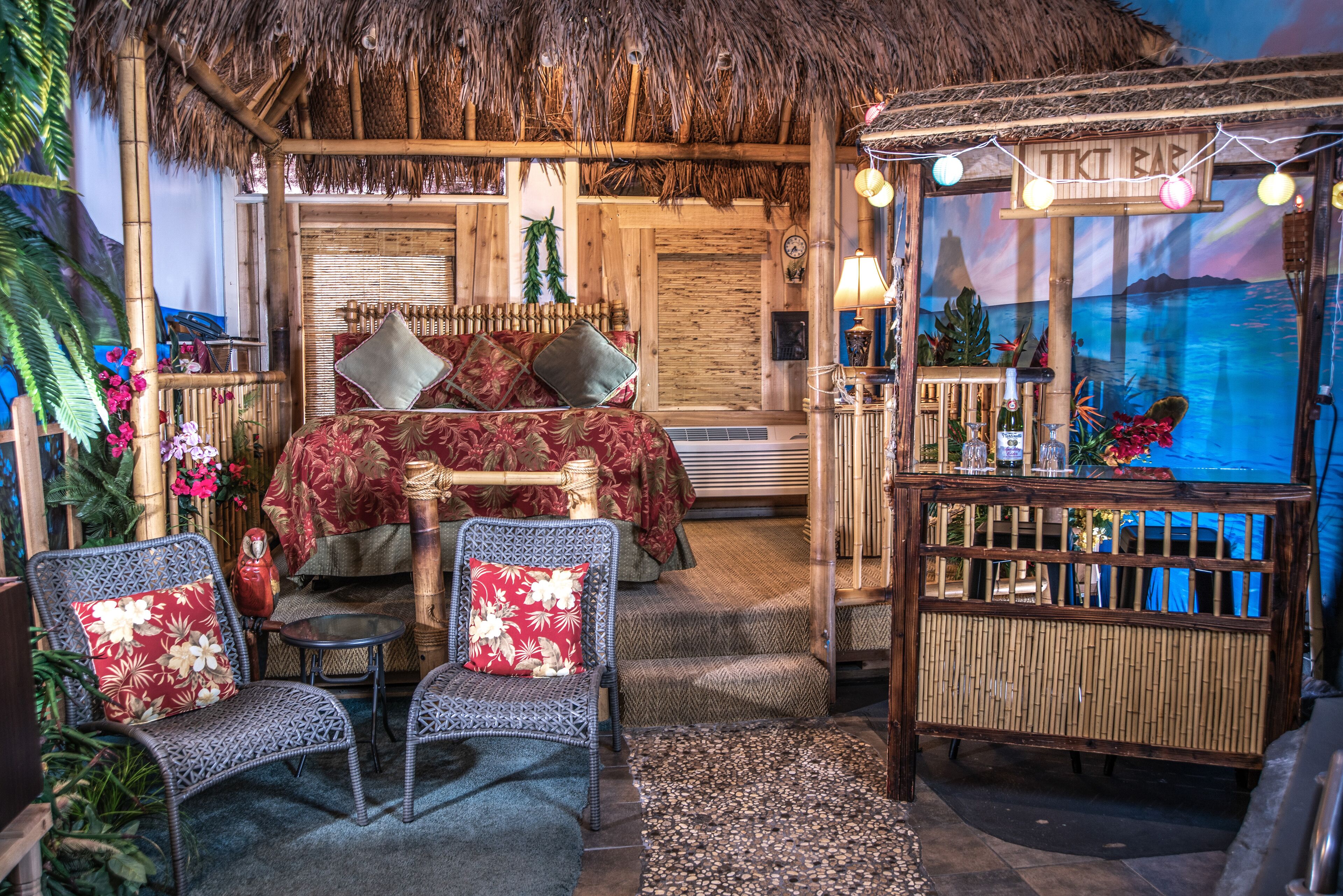 Une pièce comprenant un lit, deux fauteuils en osier, une petite table et un coin bar de style tiki.