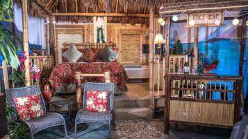 Une pièce comprenant un lit, deux fauteuils en osier, une petite table et un coin bar de style tiki.