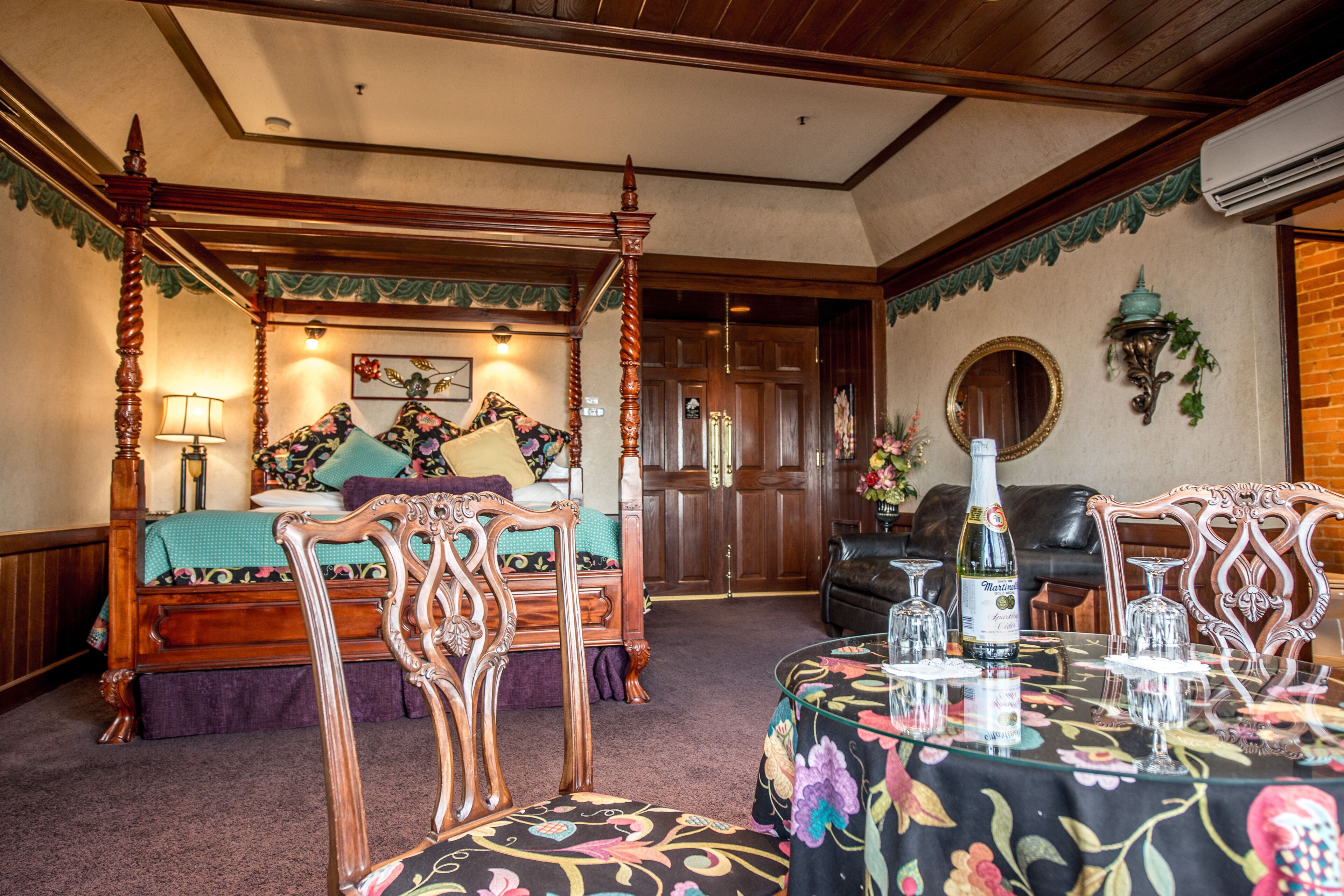 A four-poster bed with floral bedding, a sitting area with a leather sofa, and a dining table set with glassware and a bottle.