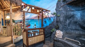 A tiki bar with a beach mural backdrop, a thatched-roof structure, and a stone wall with a towel rack. - Anniversary Inn - 5th South (Salt Lake City)