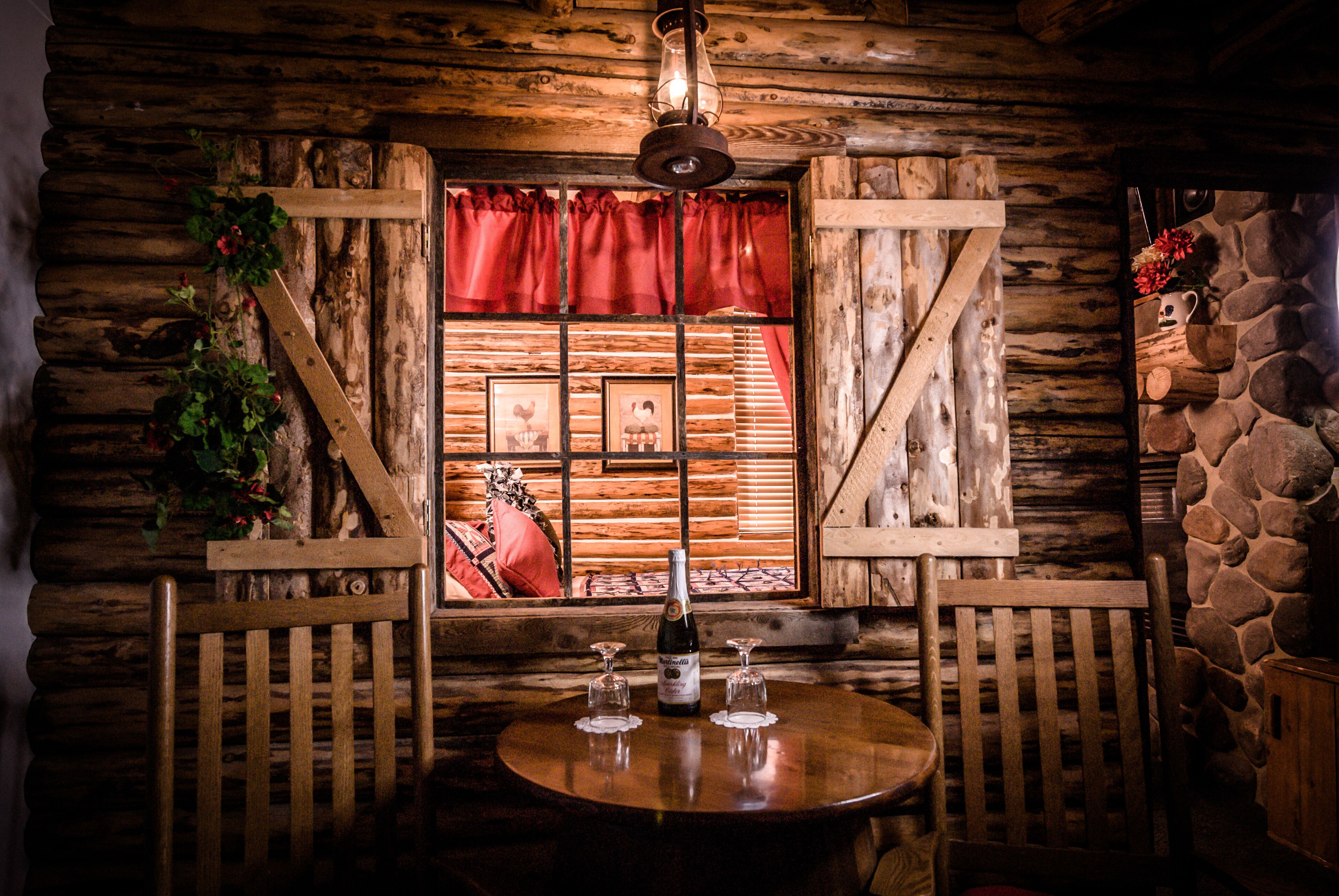 A rustic wooden cabin interior with a wooden table, two chairs, a window with red curtains, and a stone wall.