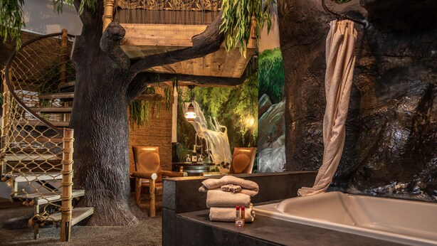 A bathroom with a large bathtub, a wooden tree structure, a hanging chair, and a waterfall mural. - Anniversary Inn - 5th South (Salt Lake City)
