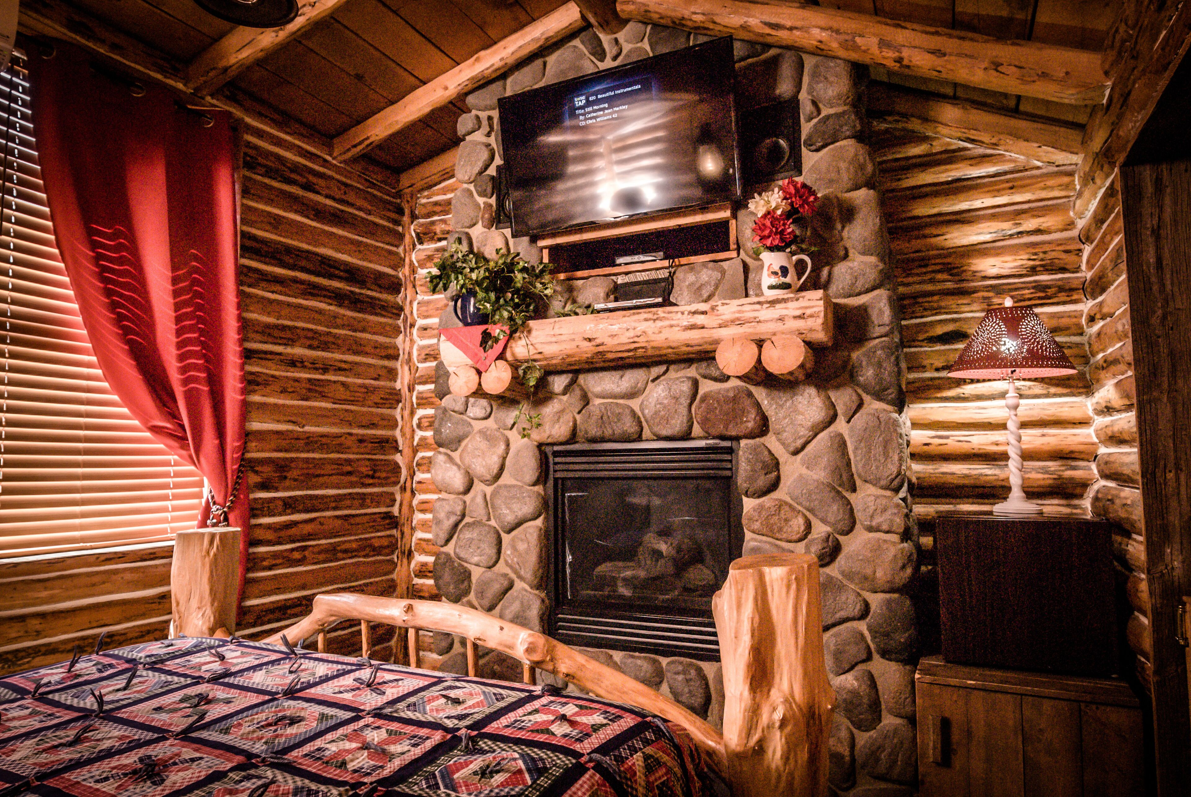 A log cabin interior with a stone fireplace, a mounted television, and a quilted bedspread.