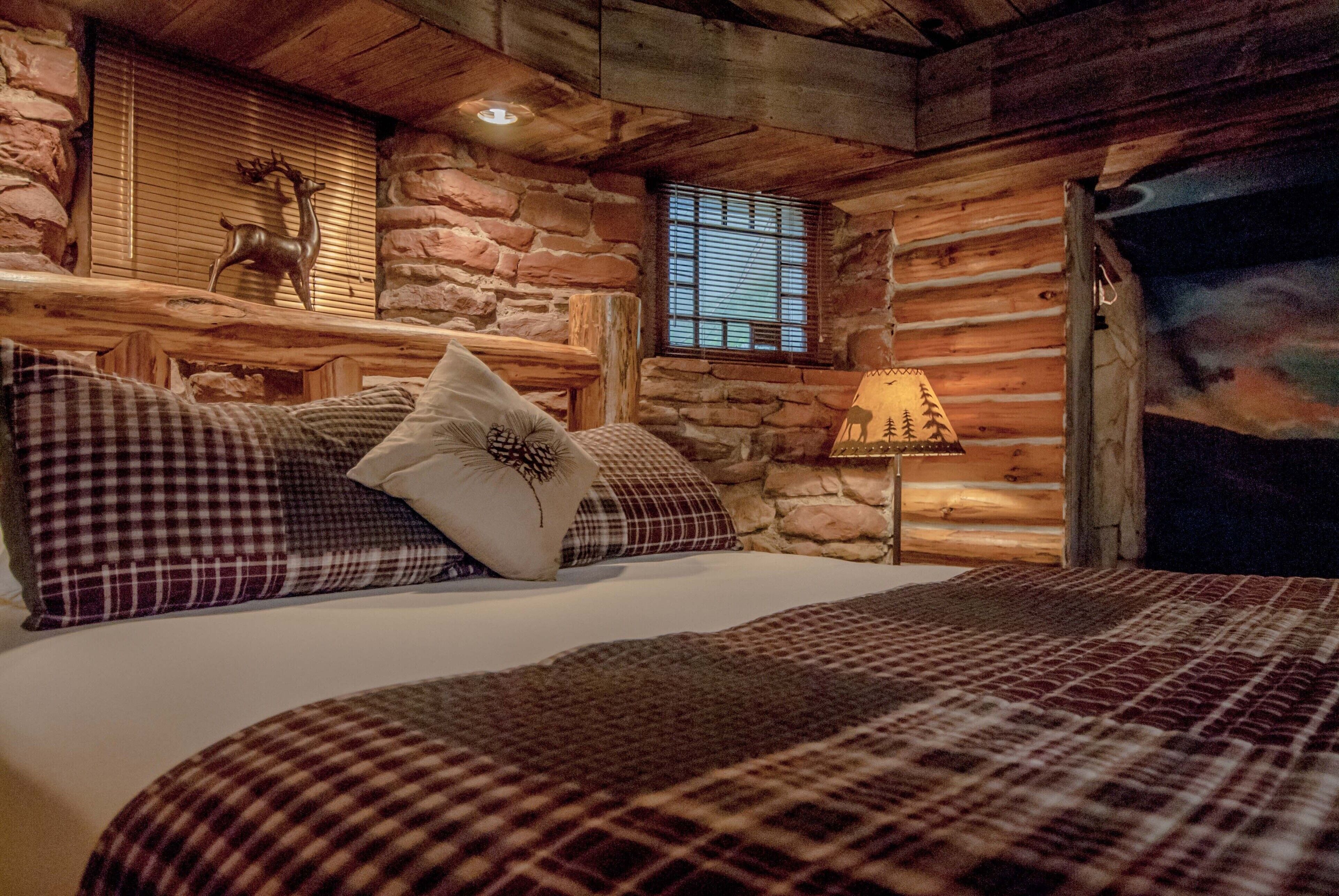 A cozy log cabin bedroom with a bed, checkered pillows, a stone wall, a window with blinds, and a lamp.