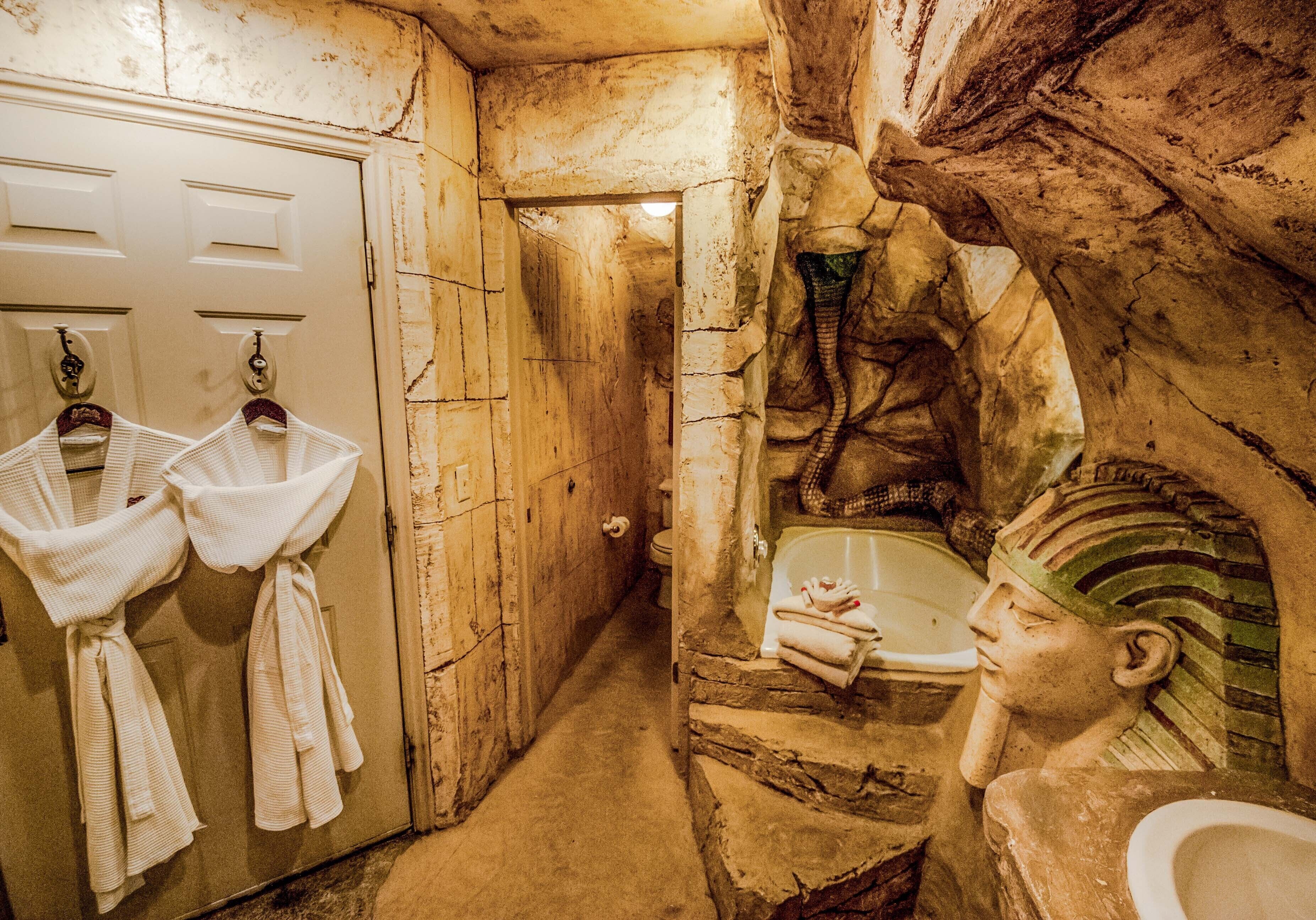 Un baño con paredes de piedra, una bañera y una estatua. Hay dos lavamanos con toallas colgadas de ganchos.