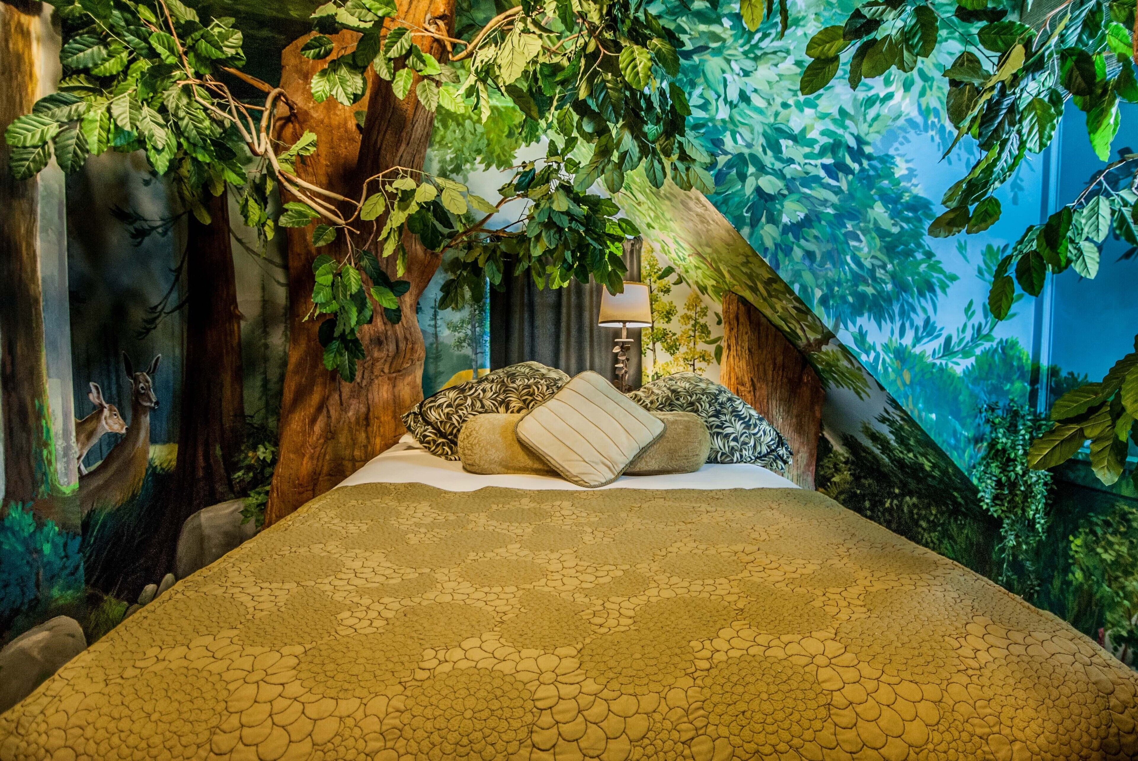 Una cama con una colcha estampada, rodeada por un mural con motivos de bosque, y una lámpara en la mesita de noche.