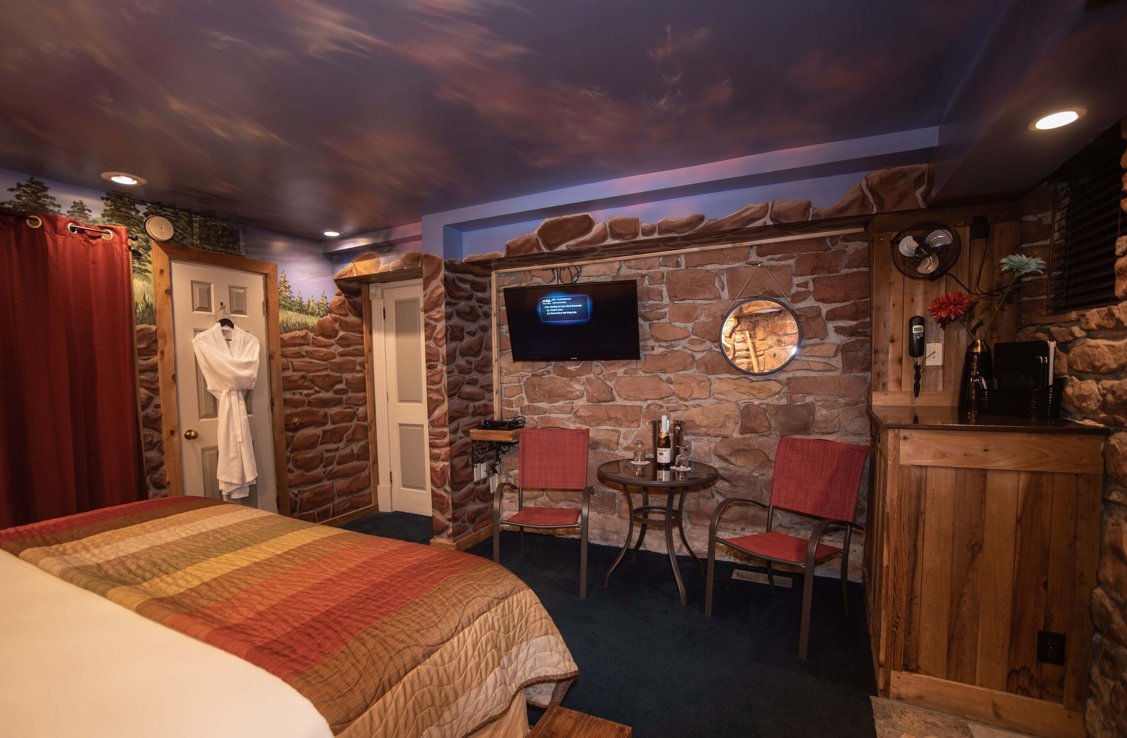 Quarto com uma cama, televisão fixada em uma parede de pedra, uma mesinha com cadeiras e um armário de madeira.