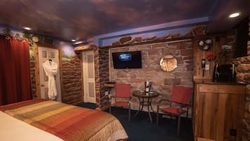 Quarto com uma cama, televisão fixada em uma parede de pedra, uma mesinha com cadeiras e um armário de madeira.