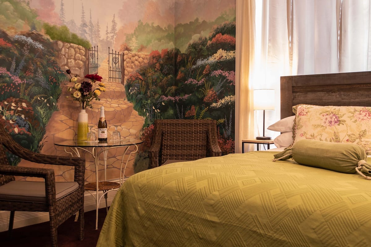 A room with a bed, wicker chairs, a small table with a bottle and glasses, and a mural of a garden scene.
