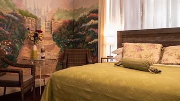 A room with a bed, wicker chairs, a small table with a bottle and glasses, and a mural of a garden scene.