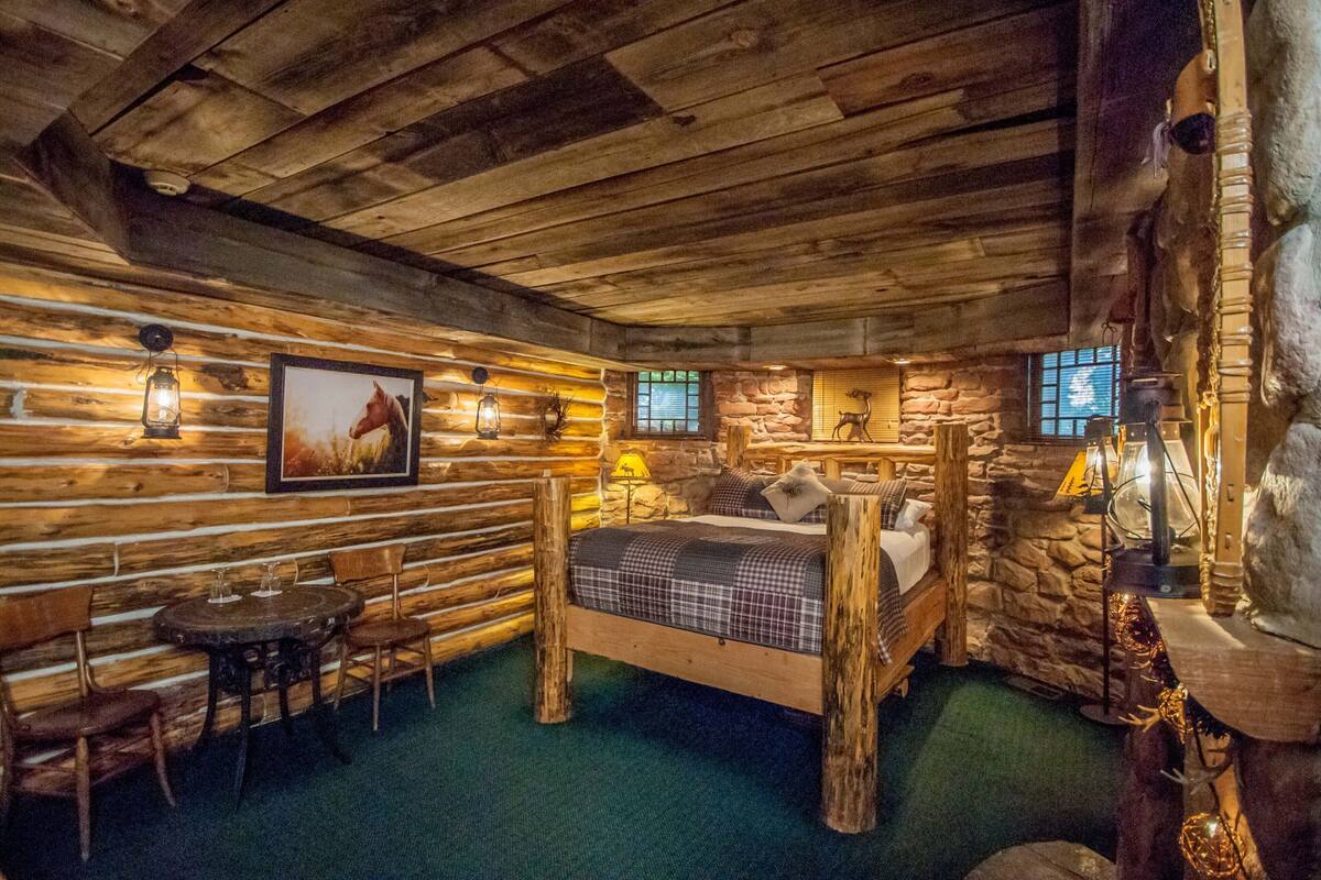 A log cabin-style room with a bed, wooden chairs, a small table, and stone walls.