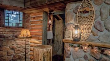 Un interior rústico con paredes de piedra, techo de madera, chimenea de piedra con un farol y una ventana con persianas.