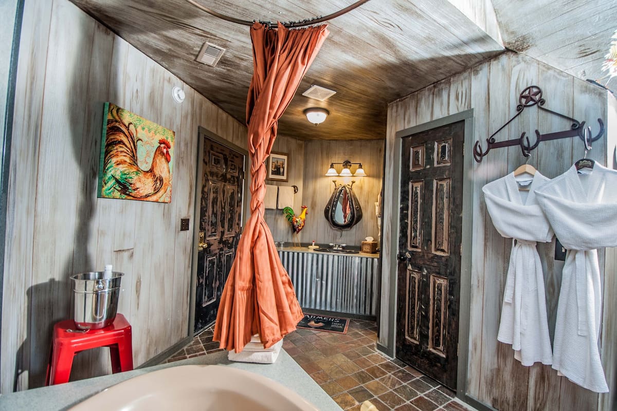 A narrow hallway with a bathtub, a red stool, a rooster painting, and a door with a towel rack.