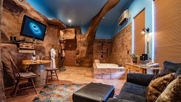 Una habitación con una gran televisión de pantalla plana, una pared de piedra, una silla de madera, un banco, un sofá, una bañera y una pequeña mesa con una lámpara.