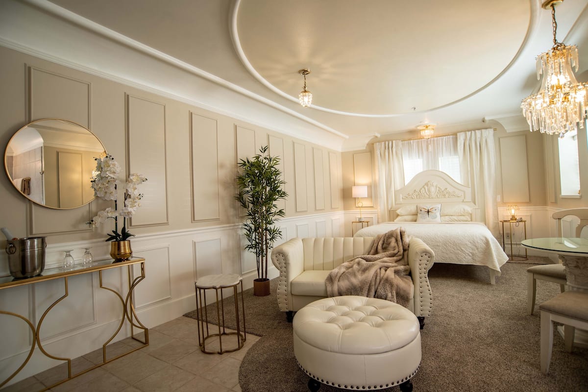Une chambre à coucher luxueuse avec un grand lit, un coin salon doté d’un pouf capitonné, une table de chevet, un miroir et un lustre.