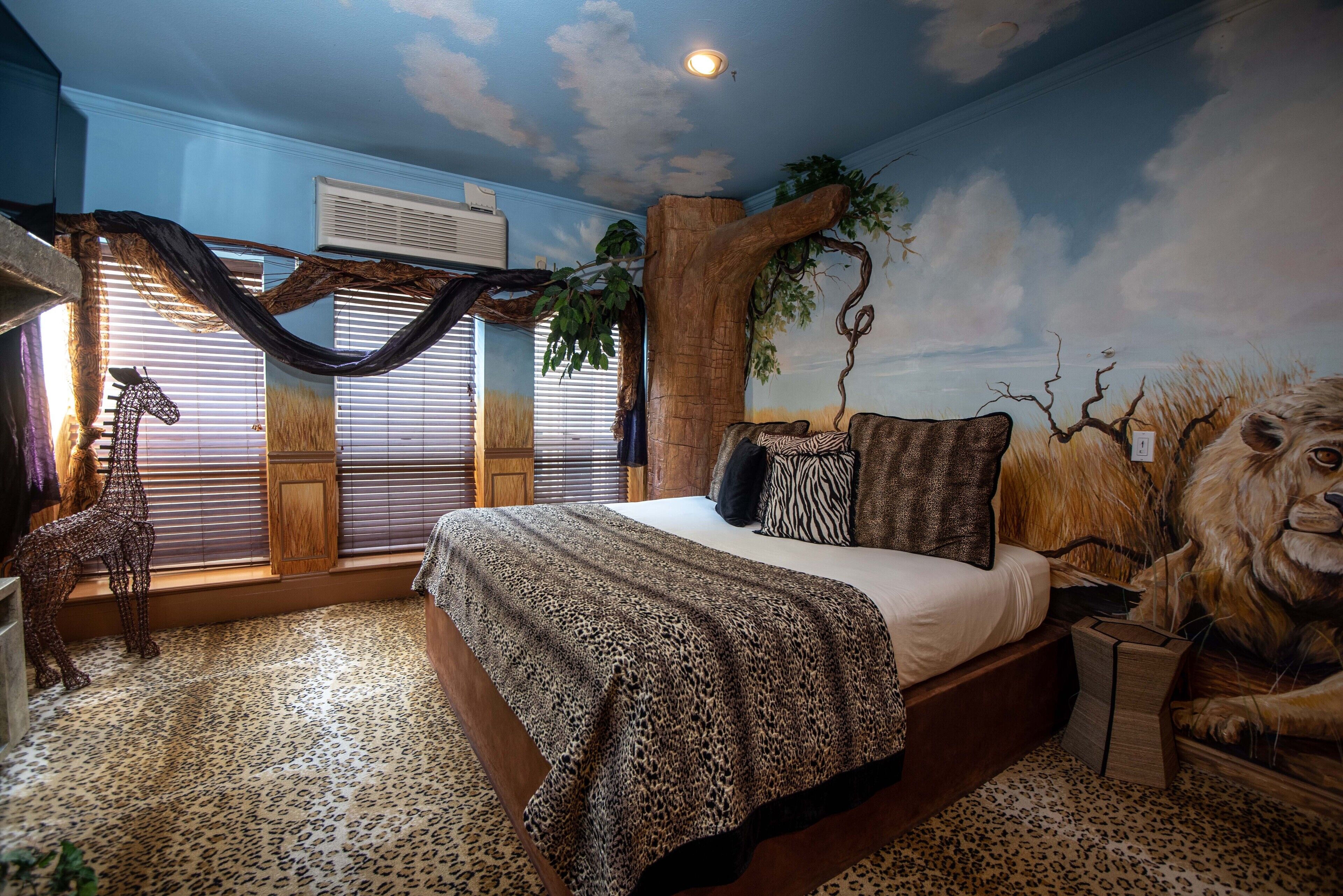 A bedroom with a bed, a large window with blinds, a tree mural, and a lion mural.