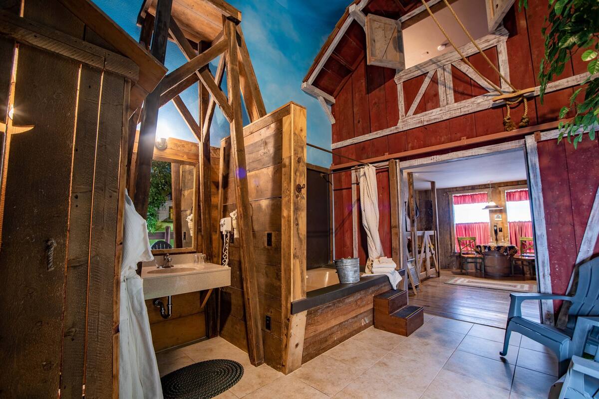 Une salle de bain rustique en bois, avec une baignoire en bois, un lavabo et un miroir. Il y a une fenêtre avec des rideaux rouges et une vue sur l’extérieur.