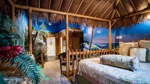 Una habitación con techo de paja, cama con ropa de cama floral y un mural con una escena tropical.