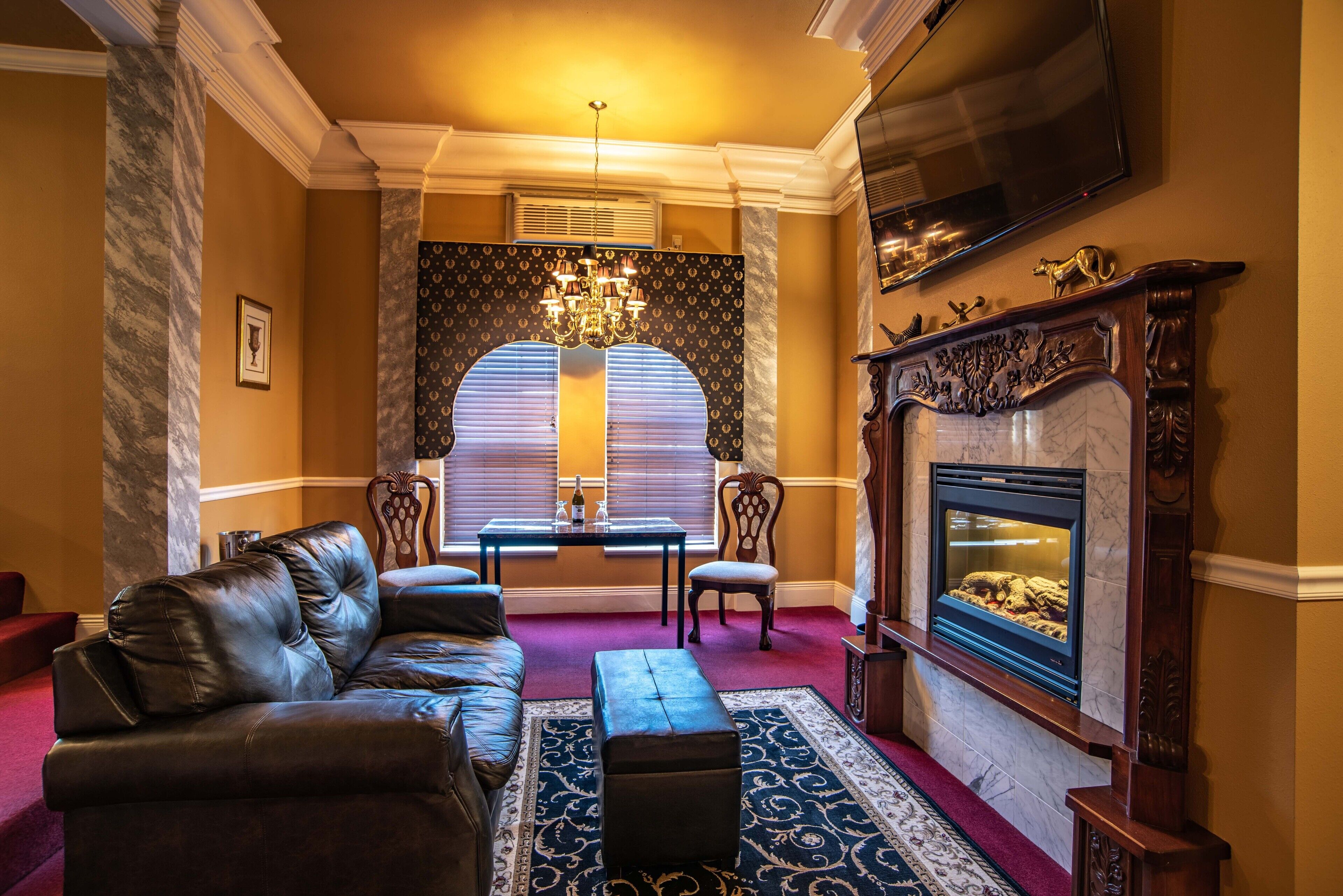 A room with a leather sofa, a small table with chairs, a fireplace, and a large window with blinds.