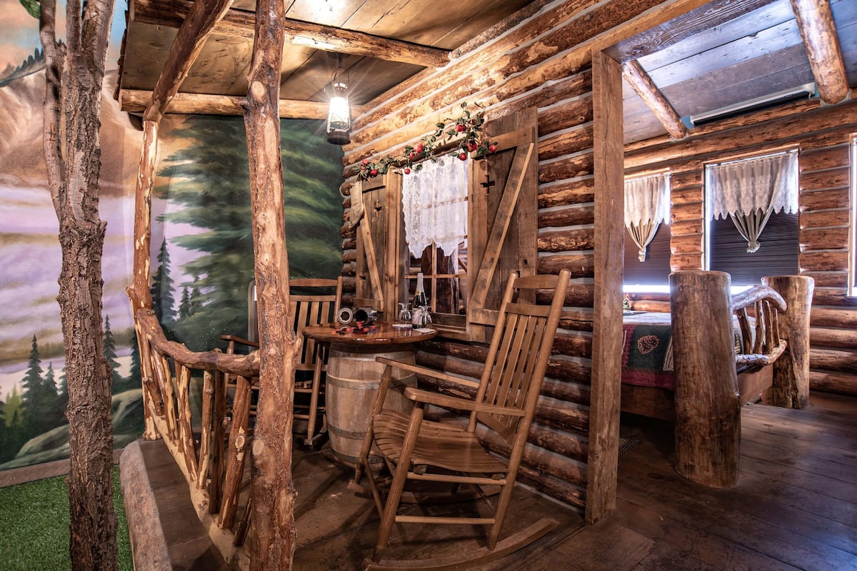 L’intérieur d’un chalet rustique en rondins de bois, avec un fauteuil à bascule en bois, une table sur laquelle est posée une lanterne et une fenêtre ornée d’un rideau en dentelle.