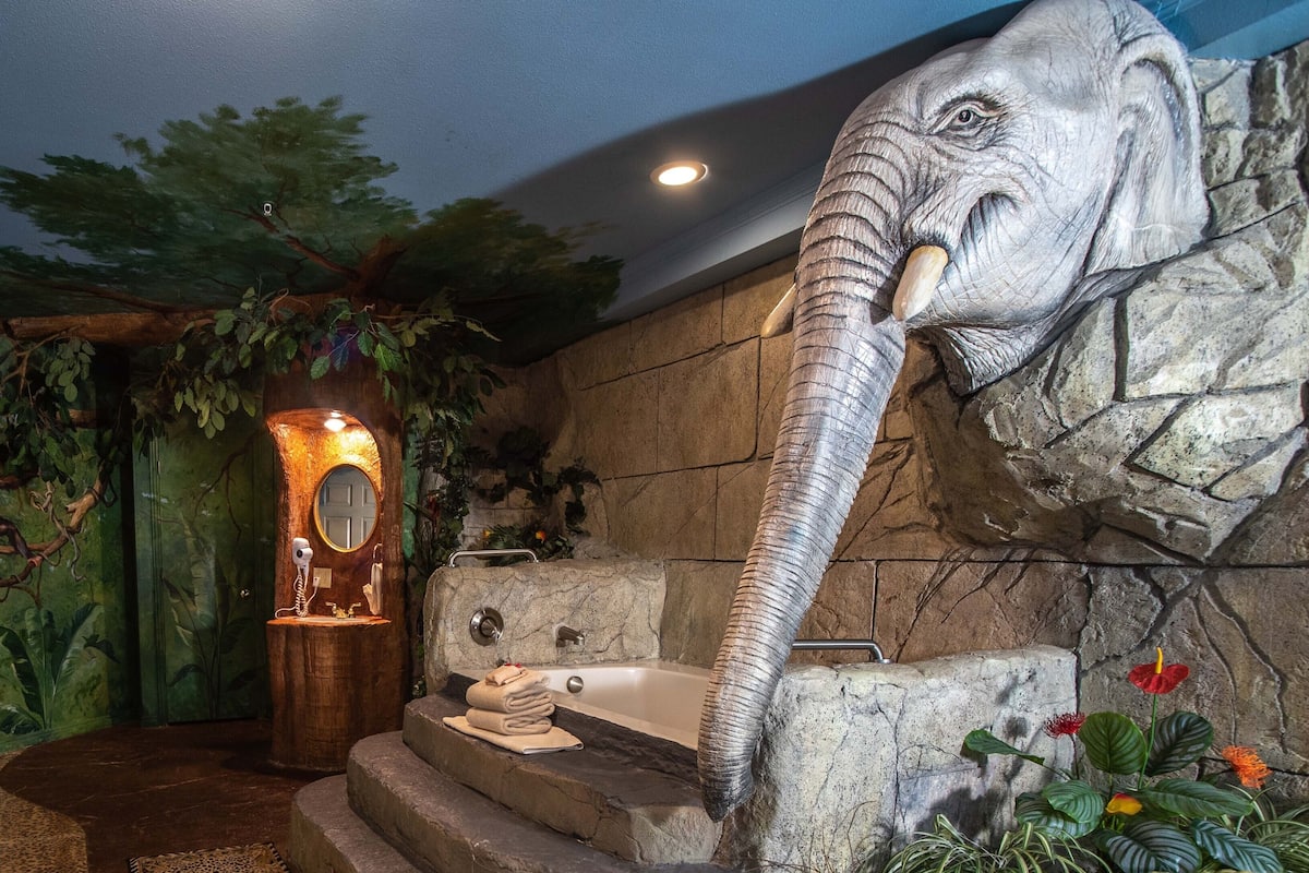 Une salle de bain avec une grande sculpture représentant une tête d’éléphant, un miroir et une baignoire.