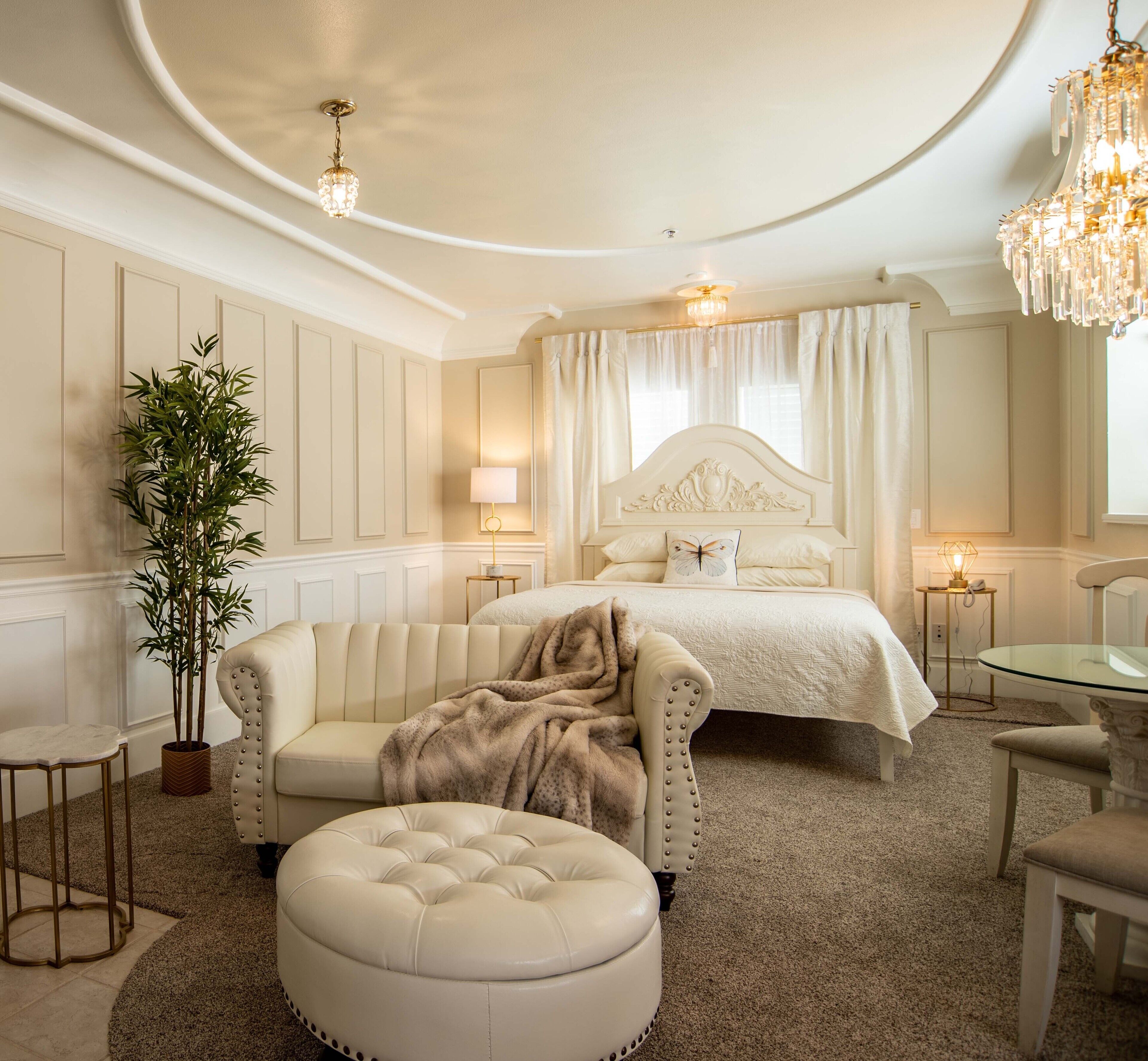 A luxurious bedroom with a large bed, a chaise lounge, a round ottoman, a potted plant, and a chandelier.