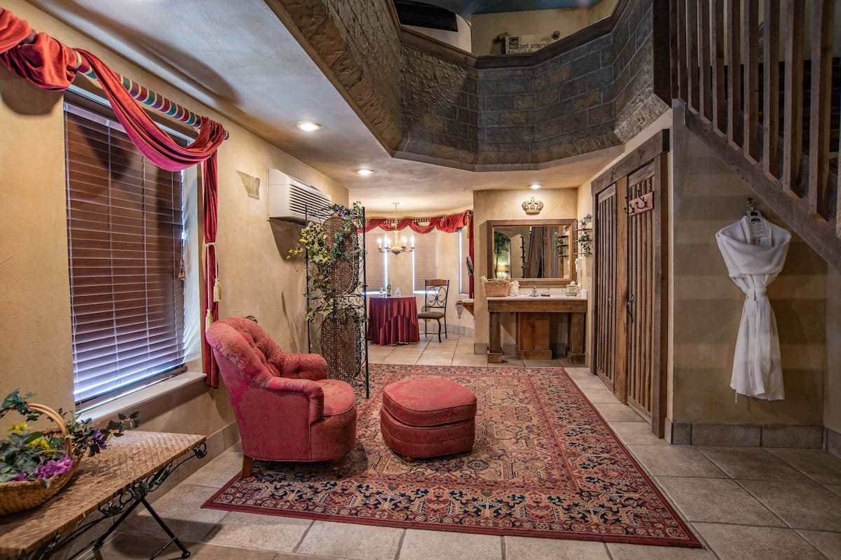 Un intérieur chaleureux avec un fauteuil rouge, un pouf et un tapis à motifs. On peut apercevoir un escalier en bois et un miroir.