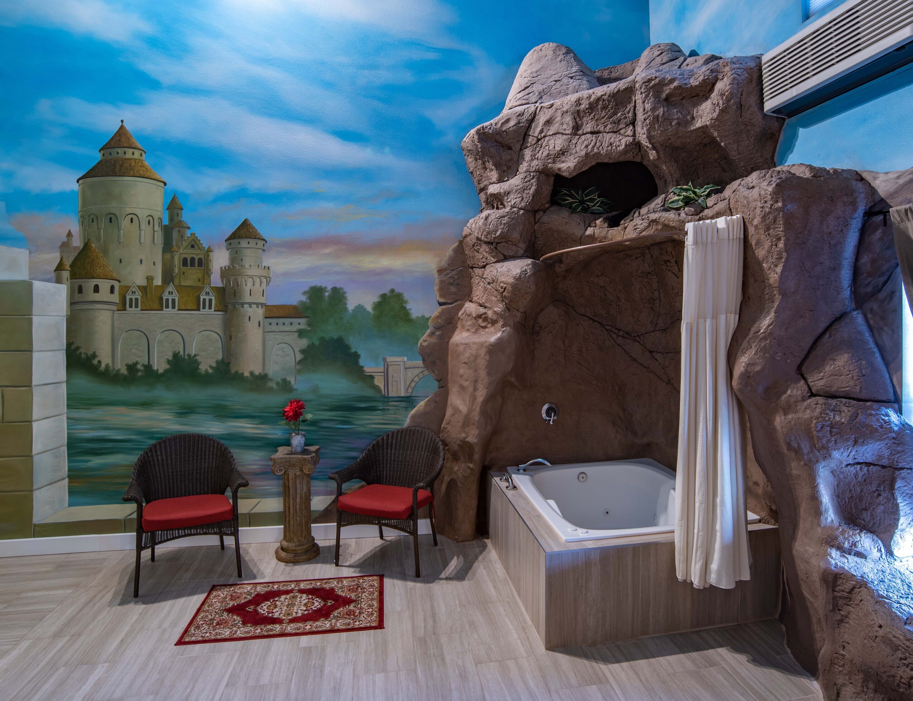 Un baño con una bañera grande, una ducha y un mural de un castillo. Hay dos sillas y una mesa con un florero de flores.