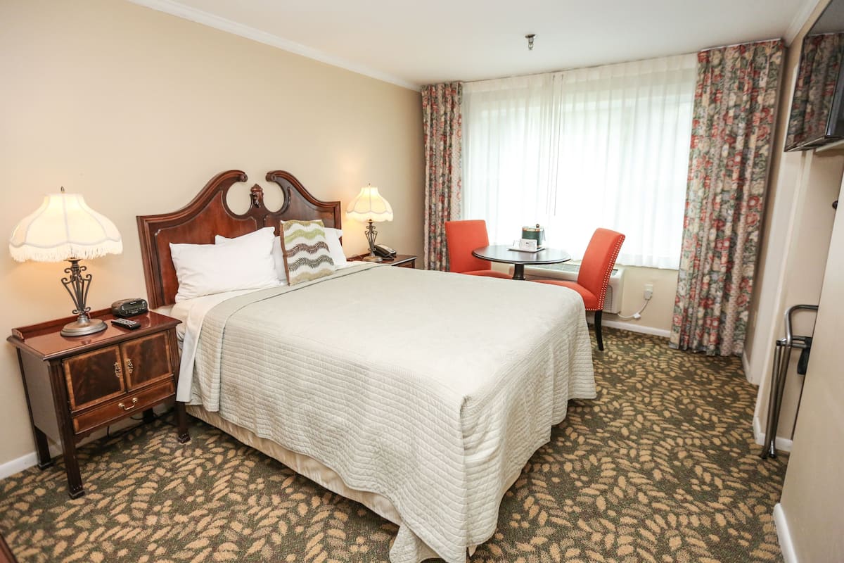 A hotel room with a large bed, two bedside tables with lamps, a small table with a chair, and a window with curtains.