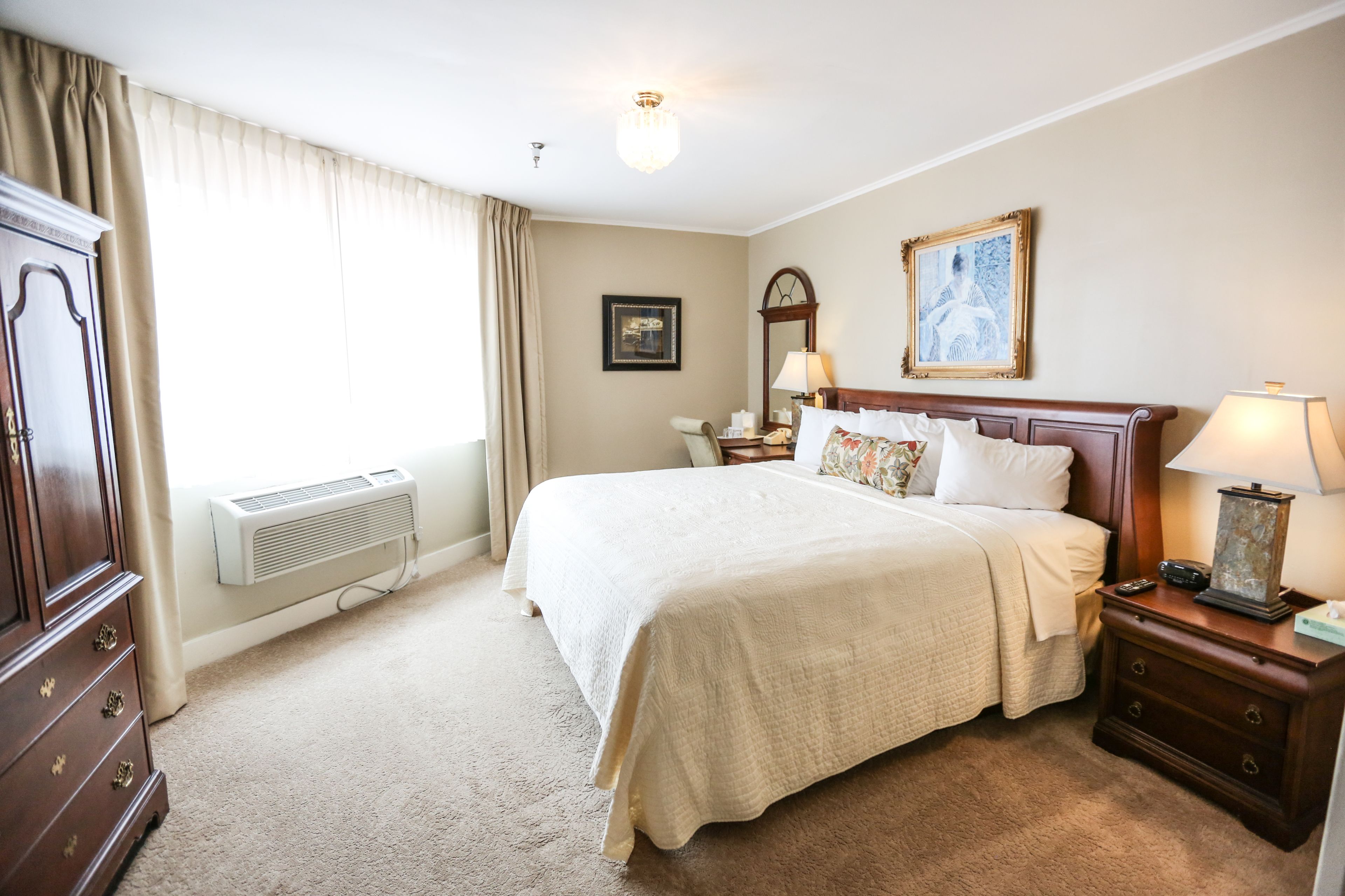 A hotel room with a large bed, a nightstand with a lamp, a dresser, a window with curtains, and a wall-mounted picture.