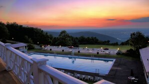 A pool with lounge chairs and umbrellas, surrounded by a white fence and trees, with a view of distant hills and a sunset.