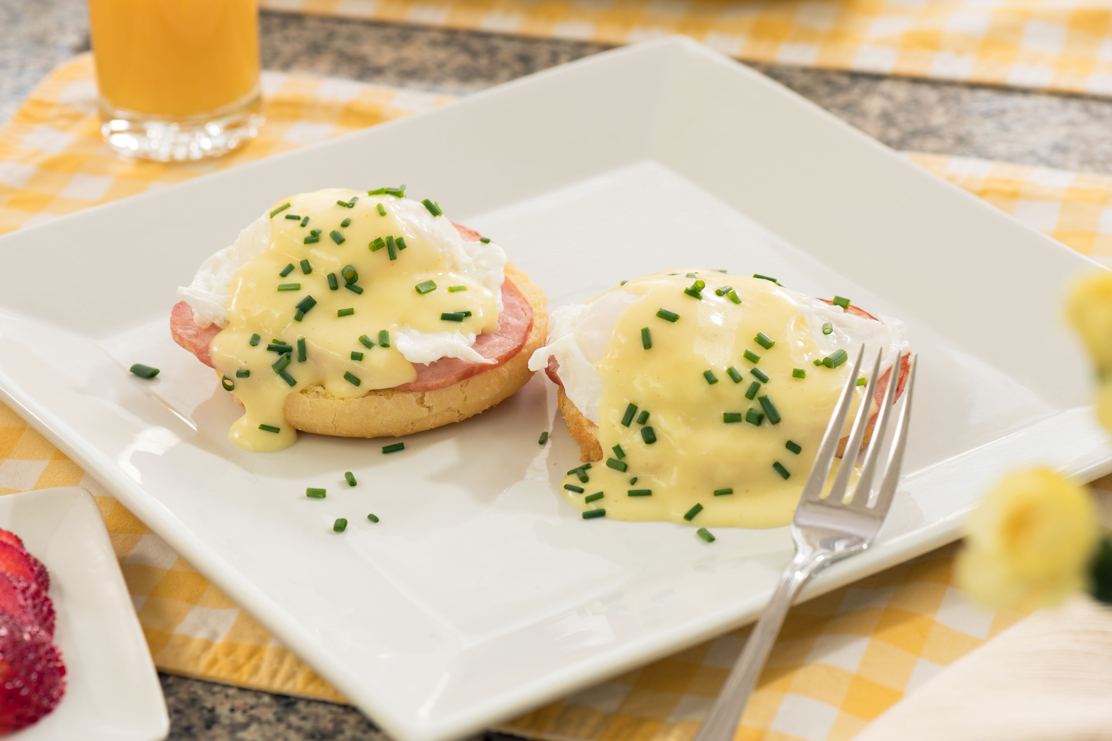 A breakfast plate with eggs benedict and a glass of orange juice on a checkered tablecloth.