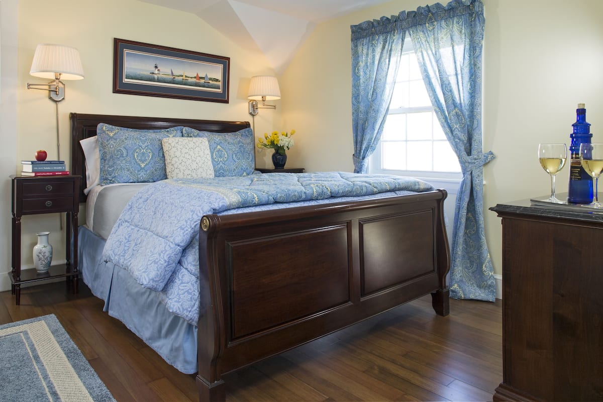 A neatly made bed with a blue and white patterned bedspread, a wooden headboard, and two bedside lamps. There is a nightstand with a vase of flowers and a stack of books. A window with blue curtains and a painting of a boat on the wall are also visible.