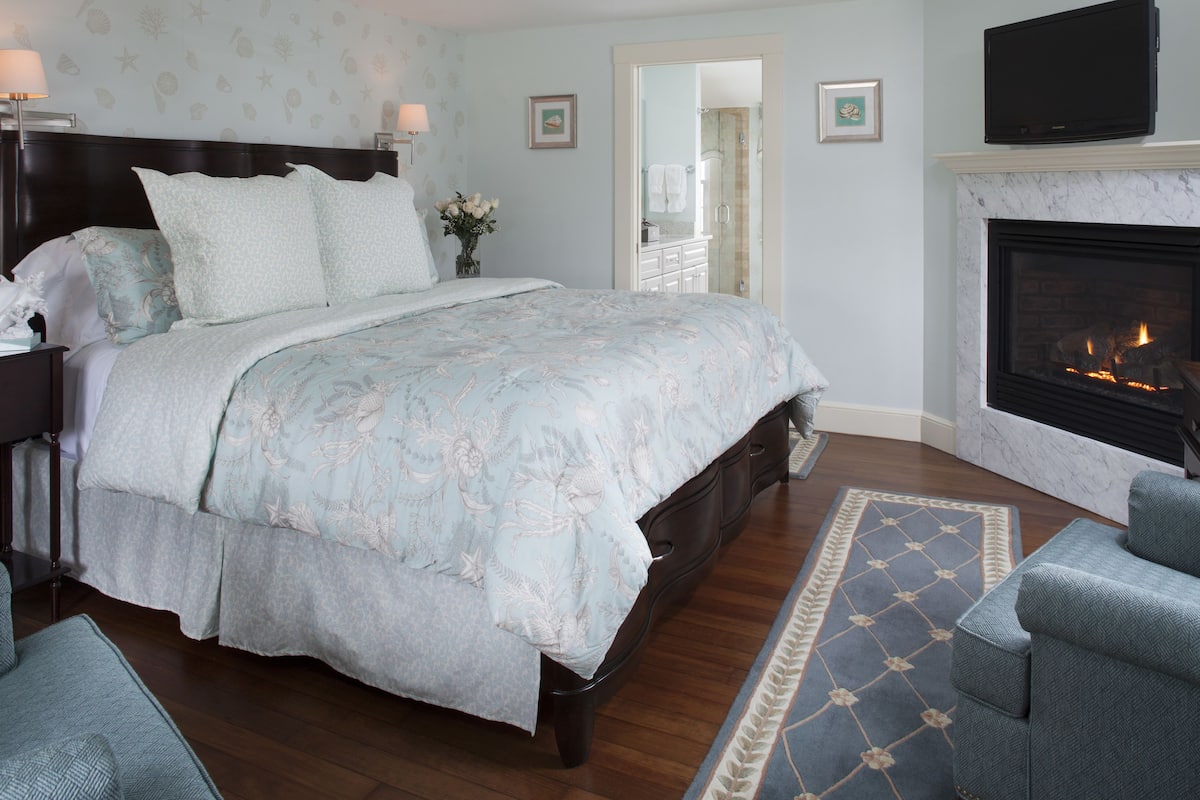 A bedroom with a large bed, a fireplace, a TV, and a bathroom visible through an open door.