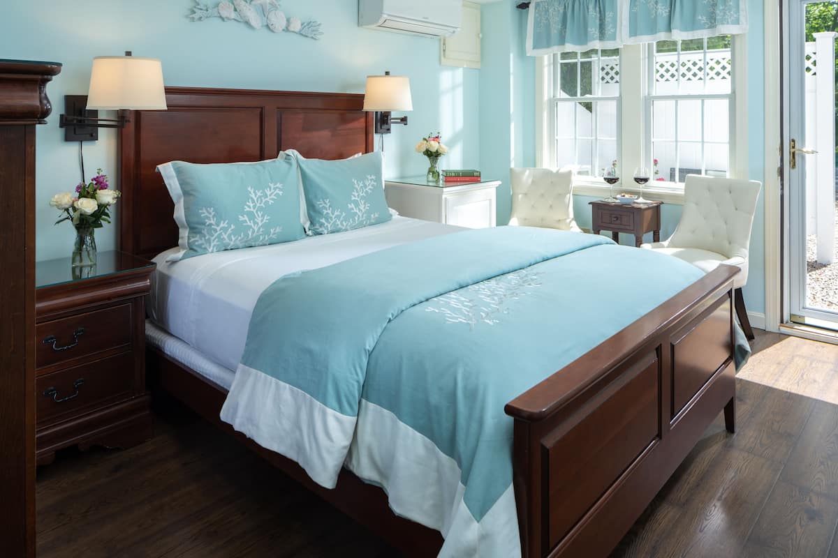 A neatly made bed with a blue and white bedspread, a wooden nightstand with a lamp, a small table with a vase of flowers, and a chair by the window.
