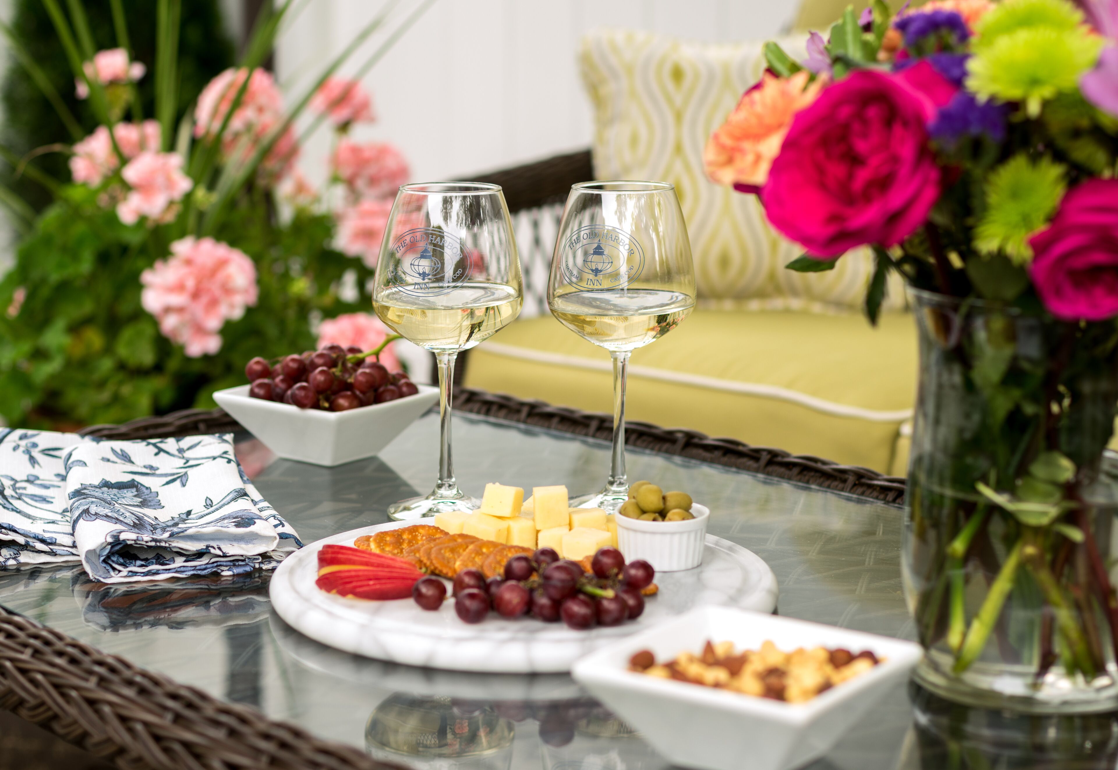 A table with a cheese platter, grapes, and two glasses of white wine, surrounded by flowers and a wicker chair.