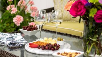 A table with a cheese platter, grapes, and two glasses of white wine, surrounded by flowers and a wicker chair.